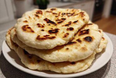 A close-up of a stack of freshly made homemade naan bread with golden-brown char marks on a white plate.
