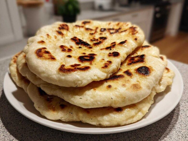 A close-up of a stack of freshly made homemade naan bread with golden-brown char marks on a white plate.