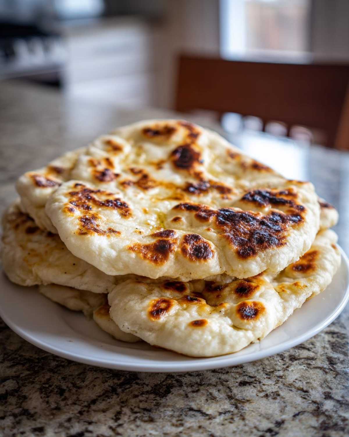 A stack of fluffy, charred homemade naan bread resting on a white plate on a granite countertop.