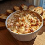 A close-up of a hot, baked reuben dip with a bubbly, browned cheese topping, served in a white ramekin surrounded by toasted bread slices.
