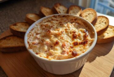 A close-up of a hot, baked reuben dip with a bubbly, browned cheese topping, served in a white ramekin surrounded by toasted bread slices.