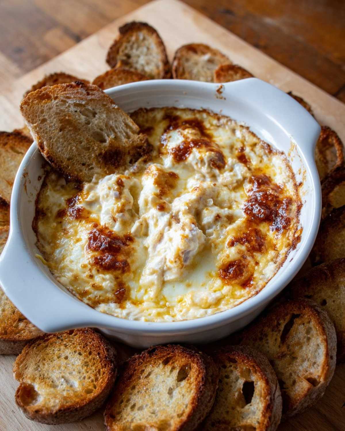 A bubbling, baked reuben dip in a white dish surrounded by toasted baguette slices for dipping.