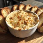 A small white bowl filled with hot, bubbly reuben dip, surrounded by toasted baguette slices on a wooden board.