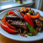 Close-up of juicy steak fajitas featuring seared steak strips mixed with vibrant red and green bell peppers and caramelized onions on a white plate.