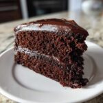 Close-up of a moist slice of dark chocolate fudge cake with glossy frosting on a white plate.