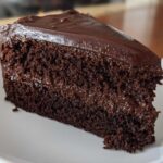 Close-up of a moist slice of dark chocolate fudge cake with rich frosting on a white plate.