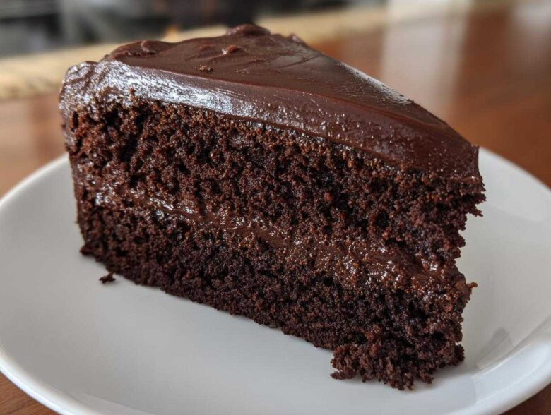 Close-up of a moist slice of dark chocolate fudge cake with rich frosting on a white plate.
