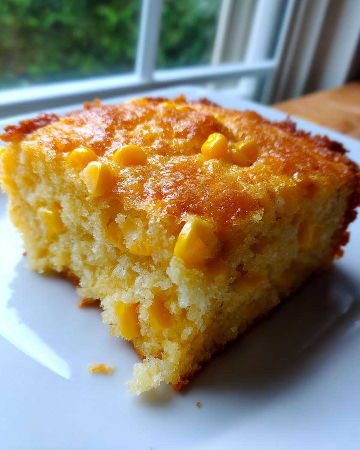 A close-up, square slice of moist cornbread casserole studded with whole yellow corn kernels, sitting on a white plate.