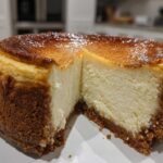 A rich, creamy slice removed from a whole new york cheesecake showing a thick graham cracker crust and a deeply caramelized top.