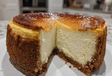 A rich, creamy slice removed from a whole new york cheesecake showing a thick graham cracker crust and a deeply caramelized top.