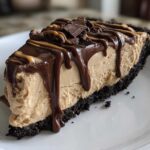 A decadent slice of no-bake peanut butter pie with a dark crust, topped with chocolate ganache and peanut butter drizzle.