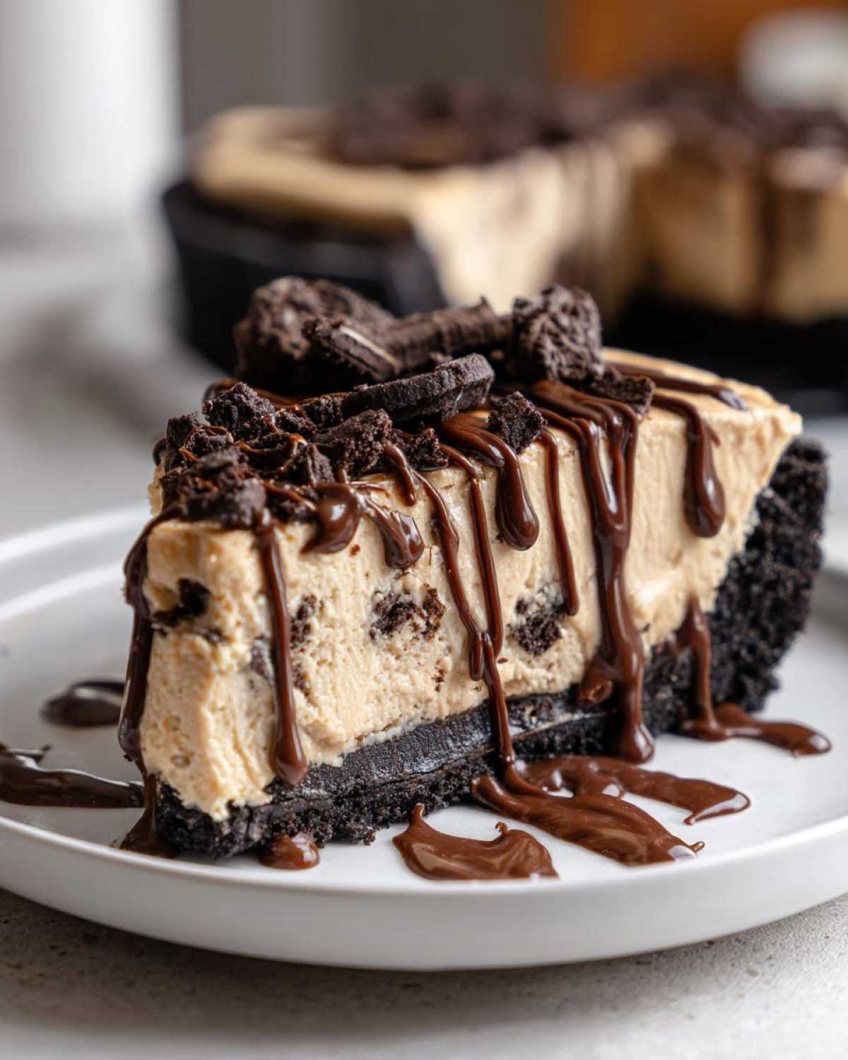 A decadent slice of no-bake peanut butter pie with an Oreo crust, topped with chocolate drizzle and cookie crumbles.
