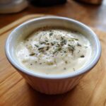 A small white ramekin filled with creamy Olive Garden Alfredo Sauce, topped with pepper and rosemary.
