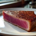 A thick, perfectly seared tuna steak, rare in the center, seasoned with spices, served on a white plate.