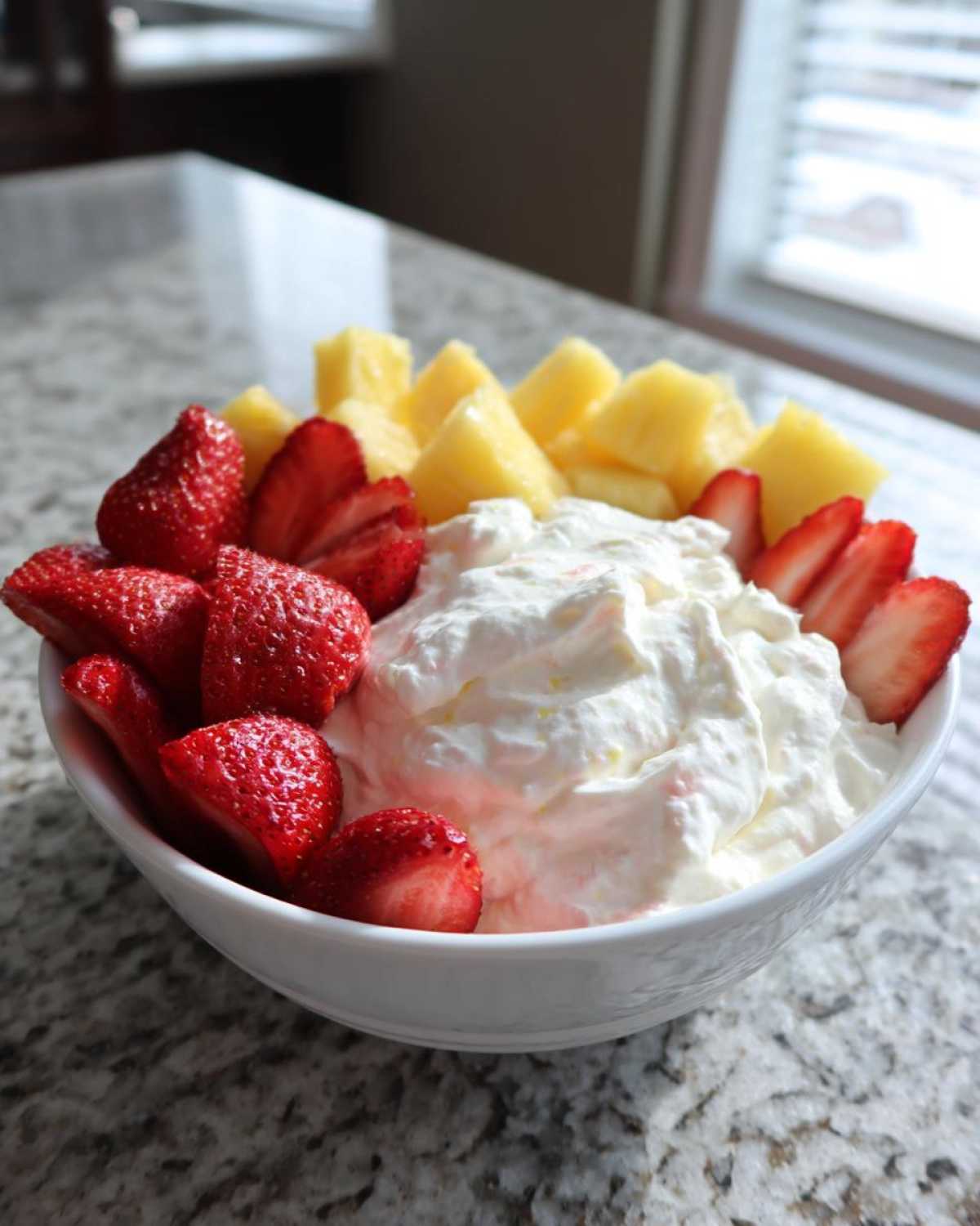 A white bowl filled with creamy fruit dip surrounded by fresh sliced strawberries and chunks of pineapple.