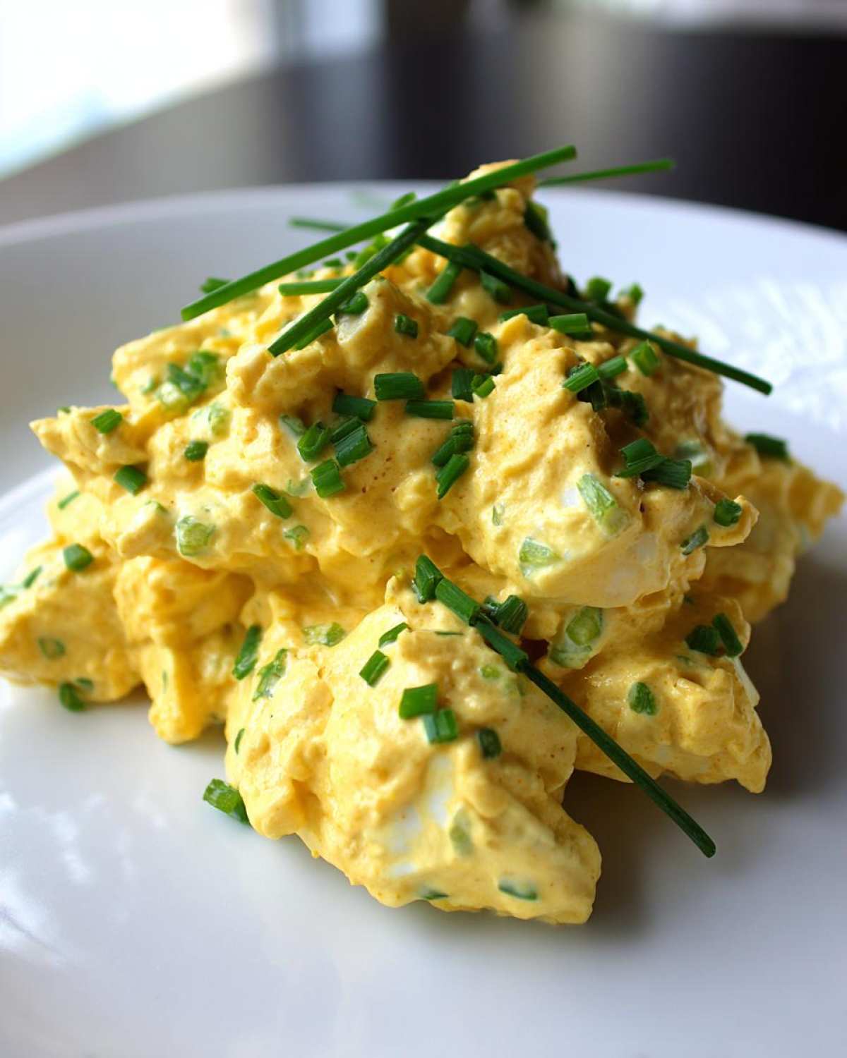 A mound of creamy, yellow egg salad recipe garnished generously with fresh, chopped chives on a white plate.
