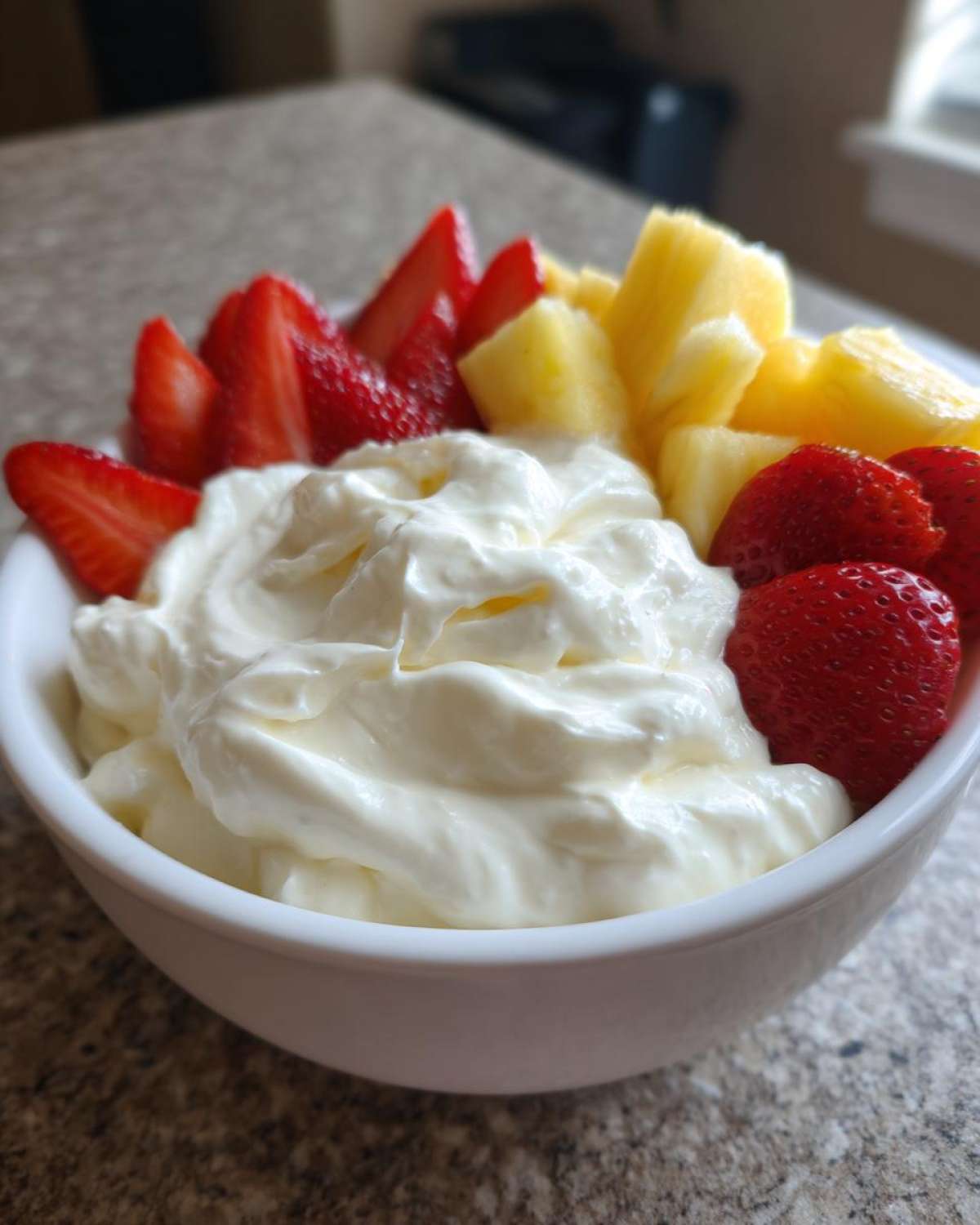 A white bowl filled with creamy fruit dip surrounded by sliced strawberries and pineapple chunks, perfect for dipping.
