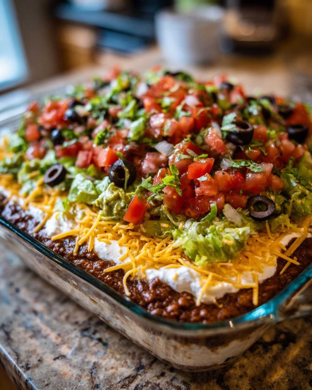 A close-up view of a fully assembled Seven layer dip in a clear glass baking dish, topped with tomatoes, olives, and lettuce.