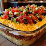 Close-up of a vibrant Seven layer dip topped with shredded cheese, tomatoes, olives, and green onions.