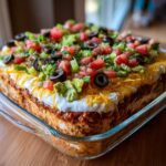 A tall, square Seven layer dip topped with shredded cheese, sour cream, tomatoes, olives, and lettuce.