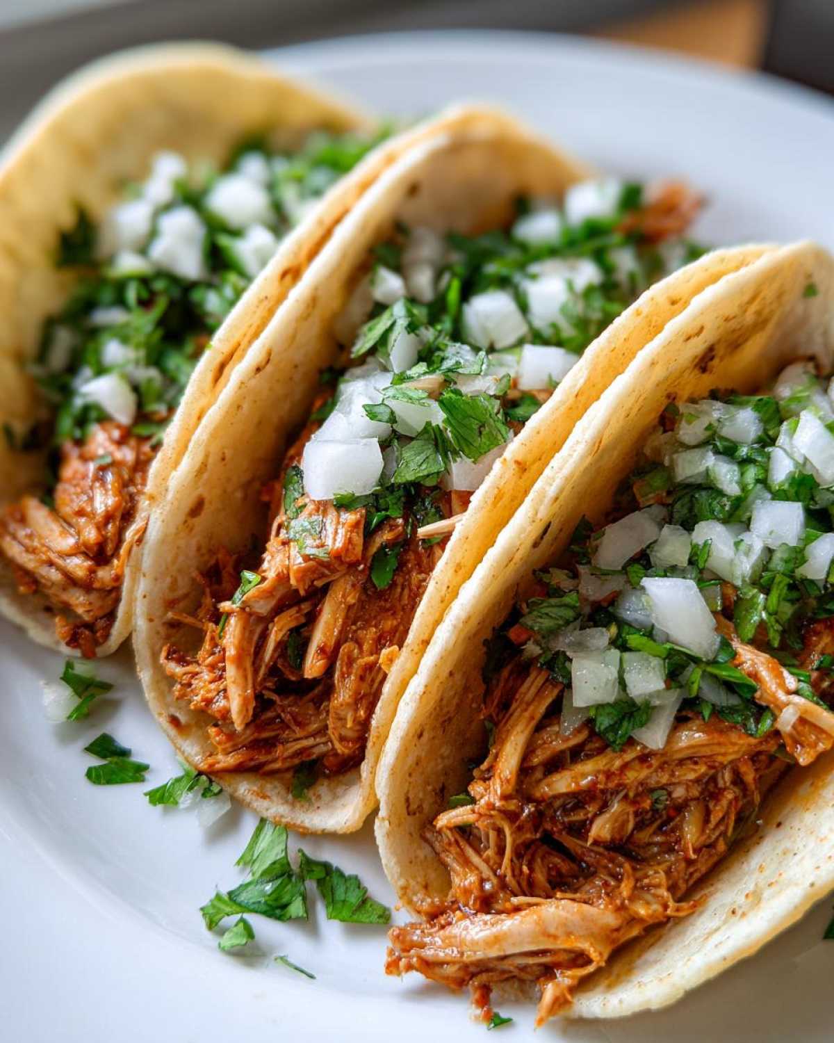 Three appetizing chicken tacos filled with shredded, saucy chicken, topped with diced white onion and cilantro.