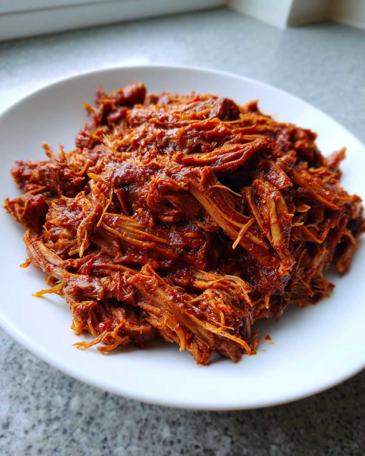 A close-up of perfectly shredded Slow cooker pulled pork coated in rich, dark red barbecue sauce on a white plate.