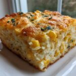 A square slice of moist cornbread casserole studded with corn kernels, featuring a golden, cheesy top garnished with parsley.