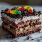 A layered slice of rich, chocolate dirt cake topped with whipped cream, crushed cookies, and colorful gummy worms.
