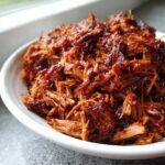 Close-up of shredded, saucy Slow cooker pulled pork piled high in a white bowl.