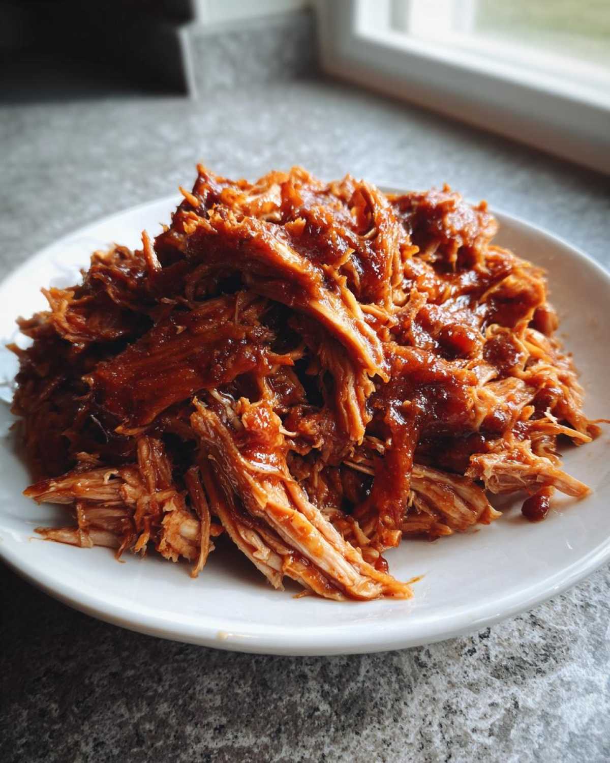 A close-up of tender, saucy Slow cooker pulled pork piled high on a white plate.