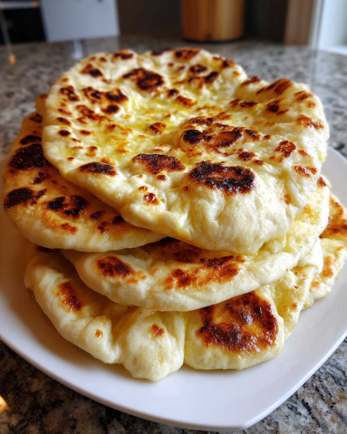 A stack of four freshly made homemade naan bread rounds with golden-brown charred bubbles on a white plate.