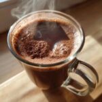 Close-up of a steaming glass mug filled with rich, dark hot cocoa mix, topped with foam and cocoa powder.