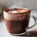 Close-up of a steaming glass mug filled with rich, dark hot cocoa mix, topped with foam and cocoa powder.