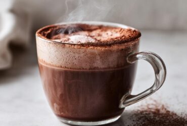 Close-up of a steaming glass mug filled with rich, dark hot cocoa mix, topped with foam and cocoa powder.