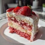 A close-up of a moist slice of strawberry cake layered with cream and topped with fresh strawberries.