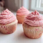 Two strawberry cupcakes with thick, swirled pink frosting sitting on a white surface in front of a bright window.