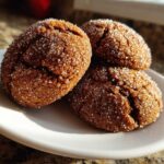 Three freshly baked gingersnap cookies, dark brown and crinkled, generously coated in sparkling white sugar, resting on a white plate.