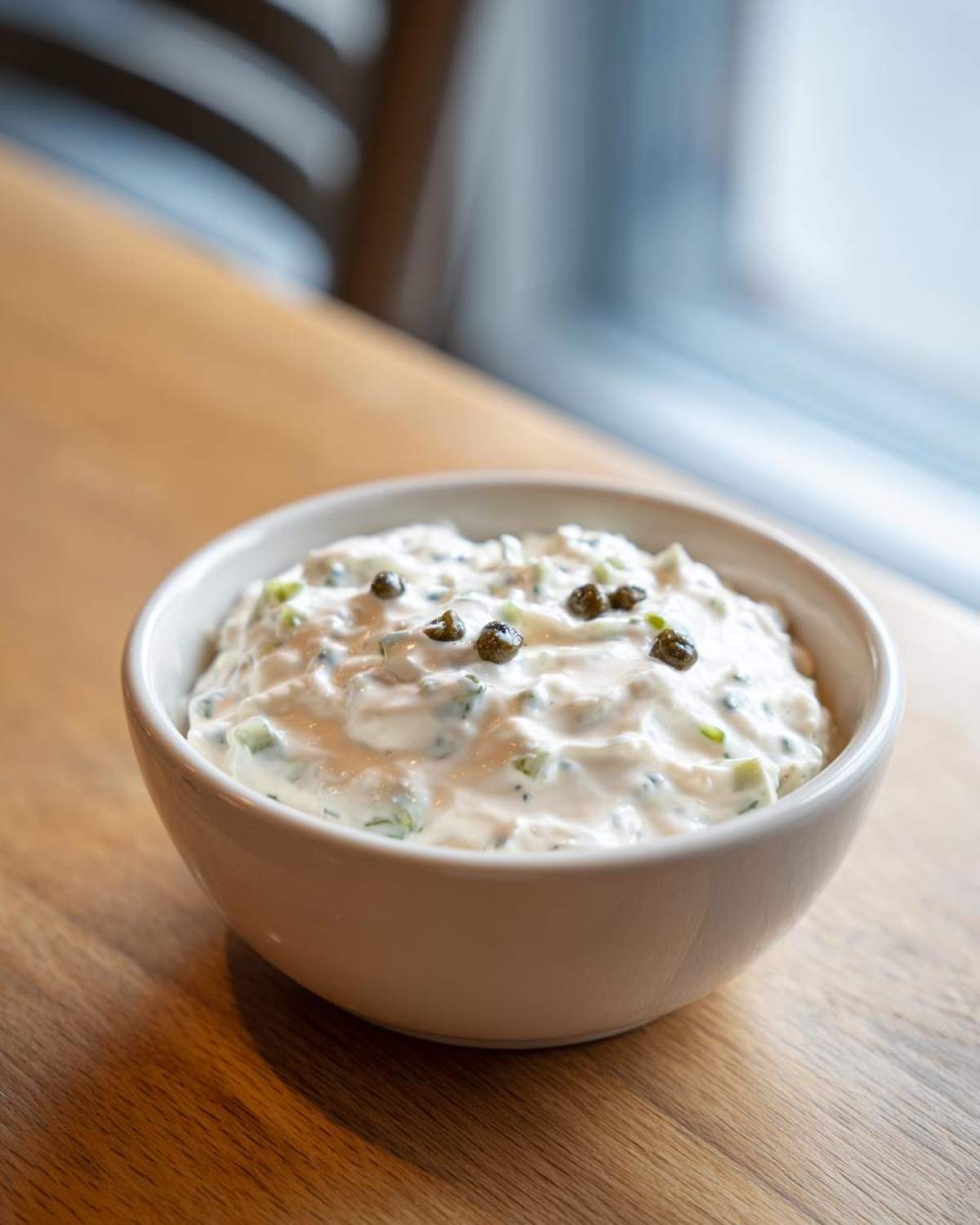 A creamy bowl of homemade tartar sauce recipe, garnished with capers, sitting on a wooden table.