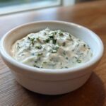 Close-up of creamy homemade tartar sauce recipe topped with fresh chopped herbs in a small white bowl.