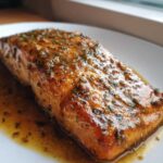 Close-up of a perfectly cooked salmon fillet glazed with a savory, herbed sauce, served on a white plate.