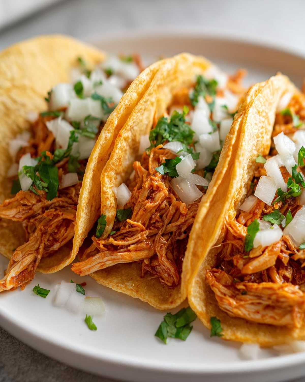 Close-up of three corn tortilla chicken tacos filled with shredded orange chicken, topped with diced white onion and fresh cilantro.
