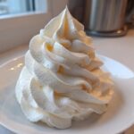 A tall, perfectly piped swirl of creamy white vanilla buttercream frosting sitting on a small white saucer.