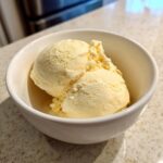 Two scoops of creamy, pale yellow protein ice cream served in a small white bowl on a speckled countertop.
