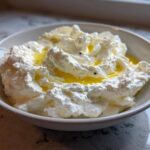 A close-up of creamy, fluffy whipped feta dip topped with golden olive oil in a white bowl.