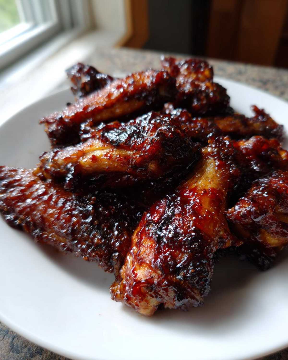 A close-up of several sticky, dark, and glossy Air Fryer BBQ Chicken Wings piled on a white plate.