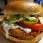 Close-up of an Air Fryer Crispy Fish Sandwich featuring a golden fried fillet, tartar sauce, tomato, lettuce, and pickles on a bun.