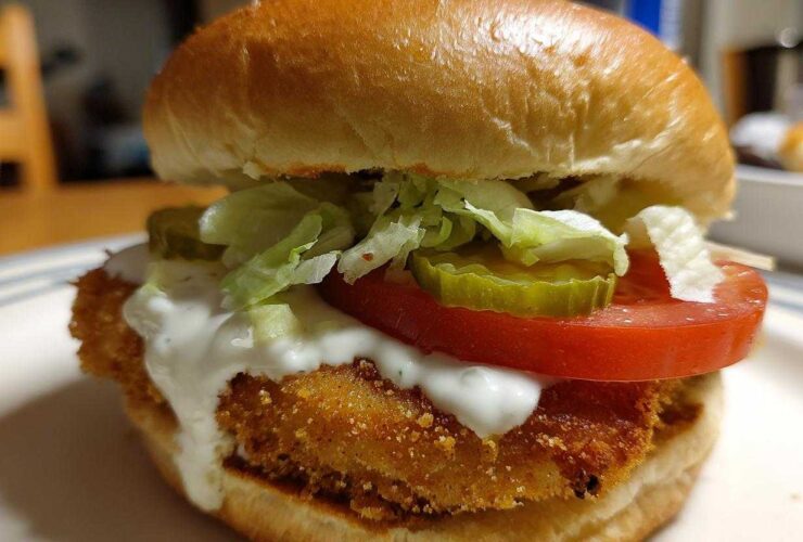 Close-up of an Air Fryer Crispy Fish Sandwich featuring a golden fried fillet, tartar sauce, tomato, lettuce, and pickles on a bun.