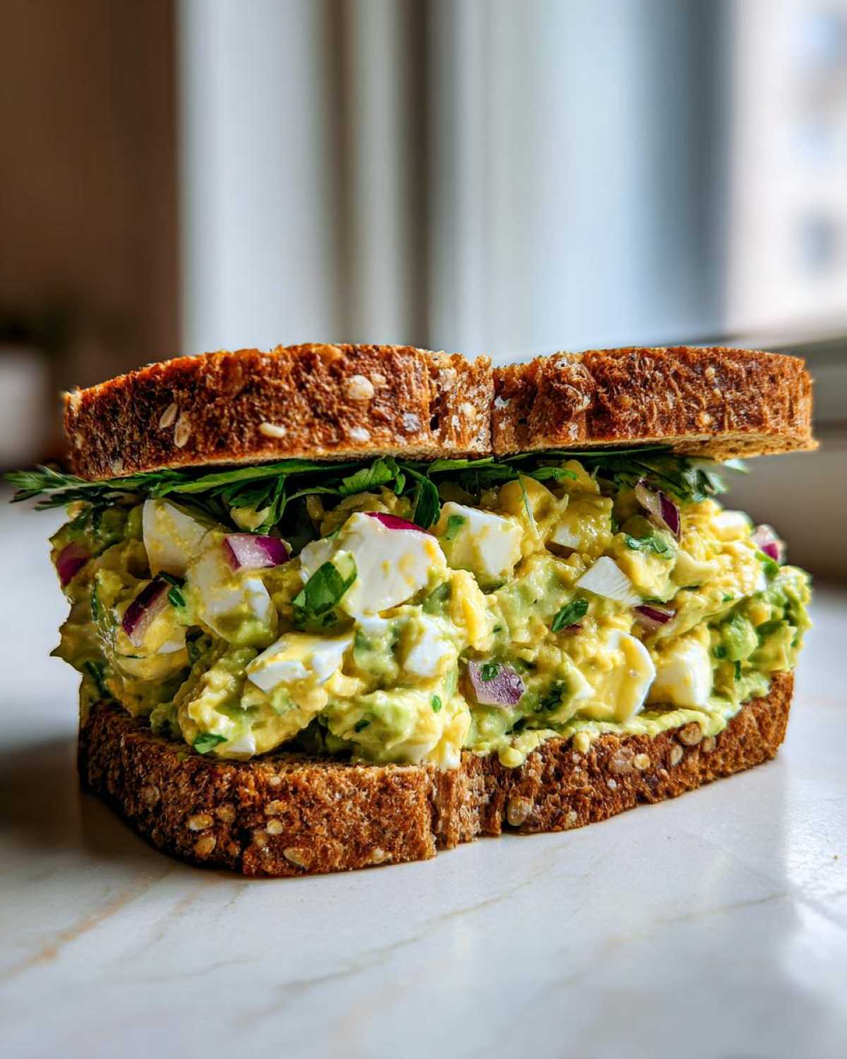 A thick slice of Avocado Egg Salad Sandwich on hearty whole grain bread, featuring chunky egg pieces and red onion.