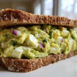 A thick, delicious Avocado Egg Salad Sandwich made with whole grain bread, visible chunks of egg white, and red onion.