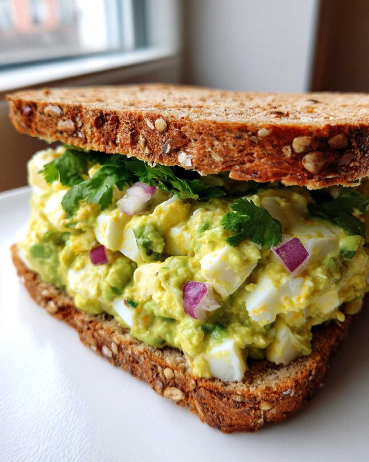 Close-up of an Avocado Egg Salad Sandwich on seeded whole grain bread with visible chunks of egg, red onion, and cilantro.